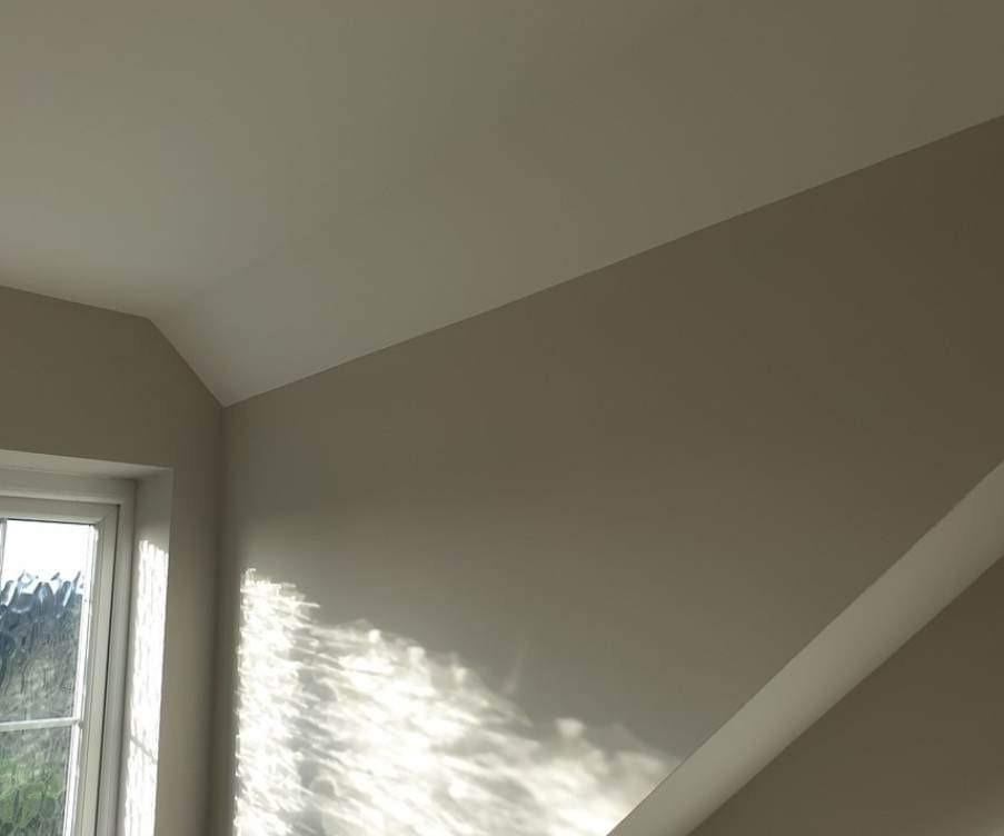 Świeżo pomalowane wnętrze z oknem, na ścianie widoczne refleksy słoneczne, gładka powierzchnia ścian i sufitu w odcieniach beżu i bieli.