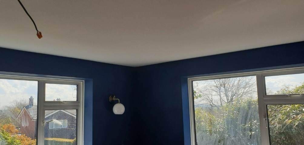 Pomieszczenie po malowaniu ścian na granatowo z białym sufitem, widoczne dwa okna z białymi ramami i widokiem na zewnątrz, na ścianie okrągła lampa, z sufitu wystaje kabel.