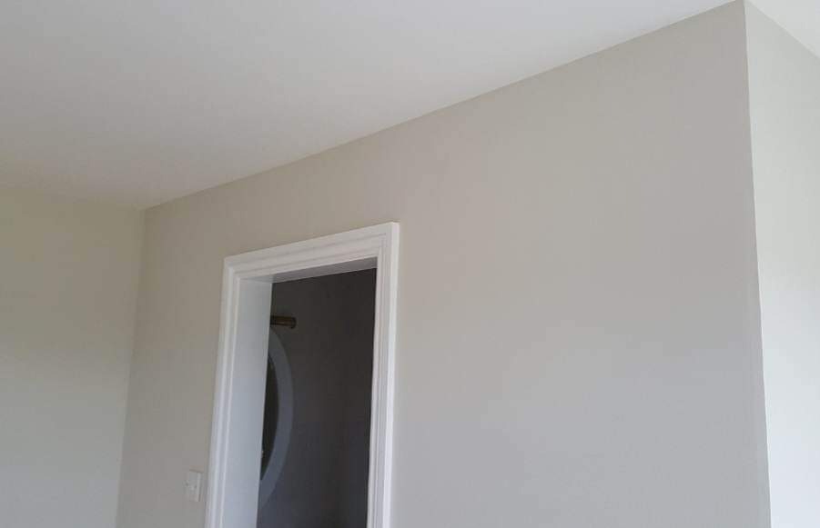 Świeżo pomalowane, jasnoszare ściany i białe obramowanie drzwi w pomieszczeniu. Widoczne delikatne cieniowanie na ścianie i suficie. Minimalistyczny wystrój wnętrza.