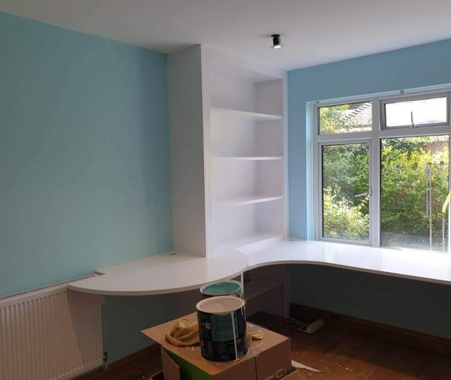 Świeżo pomalowany na błękitno pokój z wbudowanym biurkiem i regałem, widoczne puszki z farbą i wałek malarski na kartonie.