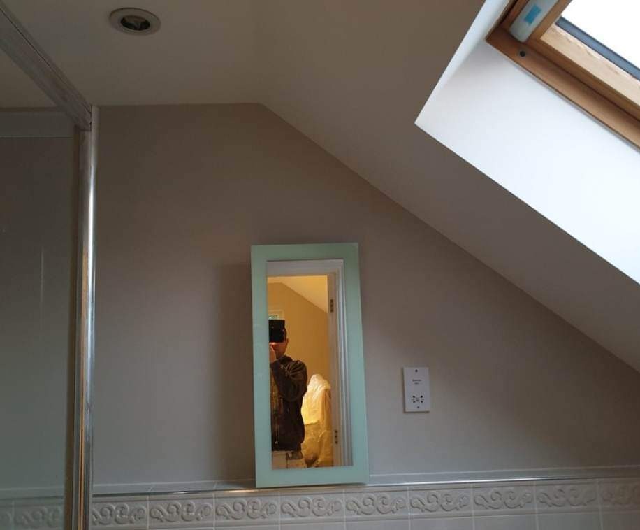 Świeżo pomalowana ściana w łazience na poddaszu, widoczny fragment kabiny prysznicowej, lustro z jasnoniebieską ramą, okno dachowe w drewnianej oprawie i gniazdko elektryczne.