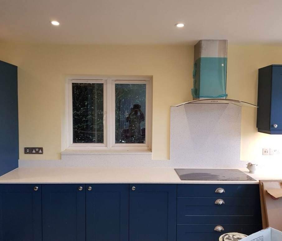 Świeżo pomalowana kuchnia z żółtymi ścianami, niebieskimi szafkami i białym blatem, okap kuchenny zabezpieczony folią ochronną, widoczne gniazdka elektryczne.