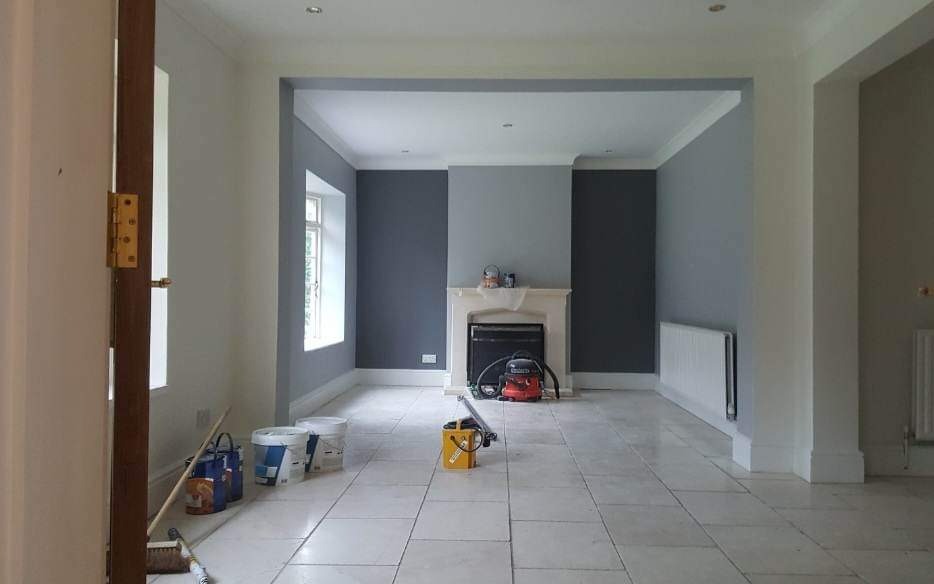 Wnętrze pokoju po odświeżeniu ścian w odcieniach szarości, widoczne puszki z farbą, odkurzacz i inne narzędzia malarskie na płytkach podłogowych.