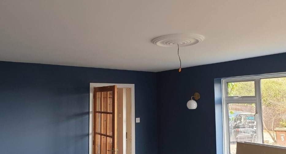 Pomieszczenie po remoncie, ściany pomalowane na granatowo, biały sufit z dekoracyjnym rozetą, drewniane drzwi z szybkami, widok na zewnątrz przez okno.