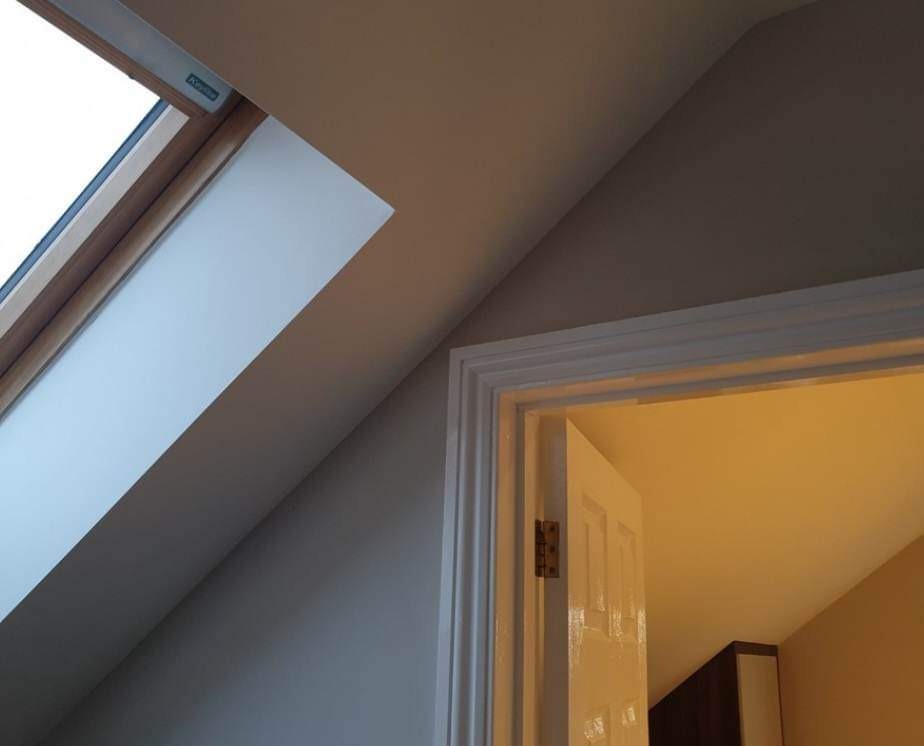 Świeżo pomalowane poddasze z oknem dachowym i otwartymi drzwiami, widoczne zawiasy, ściany w odcieniach beżu i szarości.