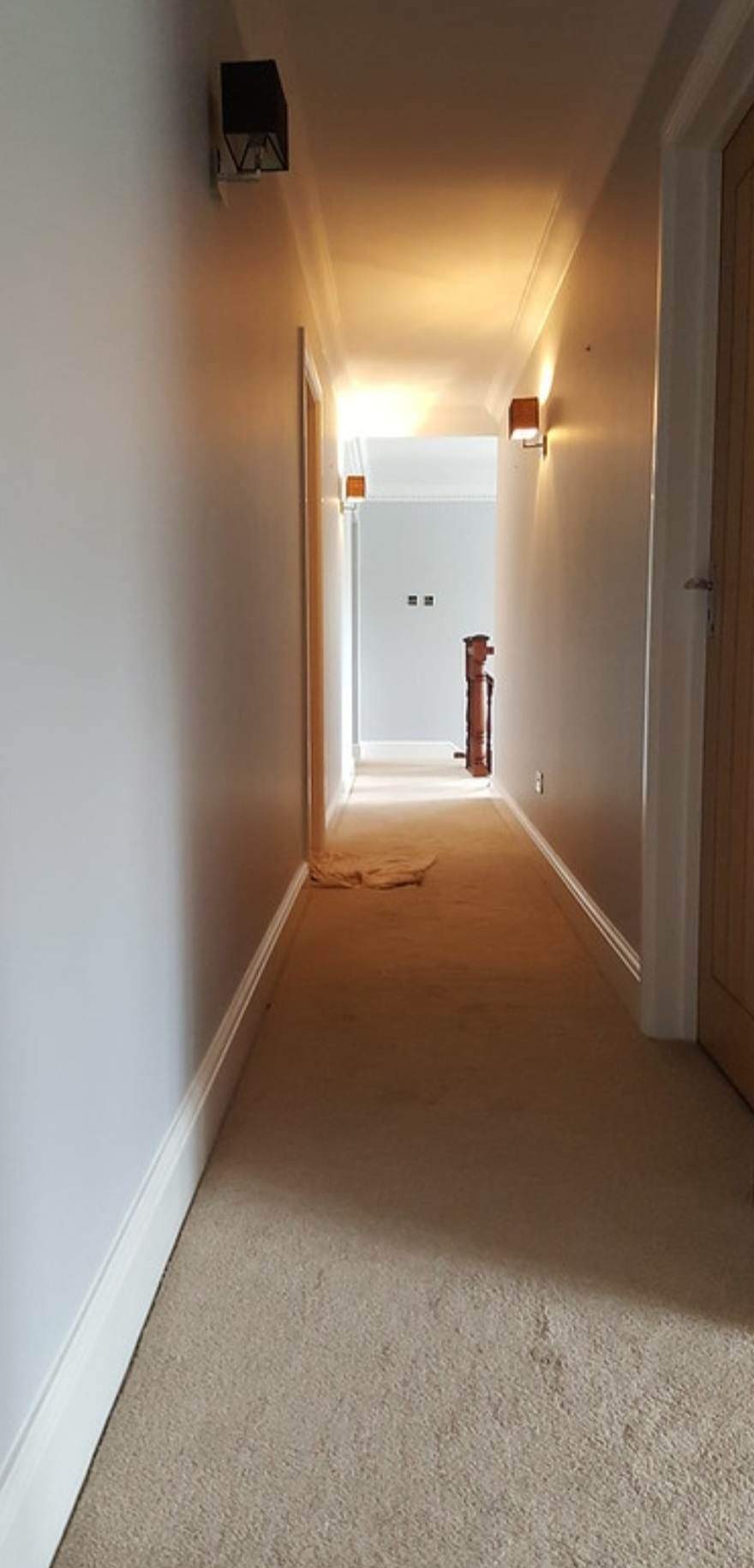 Długi korytarz po remoncie, ściany pomalowane na jasny szary kolor, widoczne drewniane drzwi i lampy ścienne, na podłodze beżowa wykładzina, zabezpieczona fragmentem materiału.