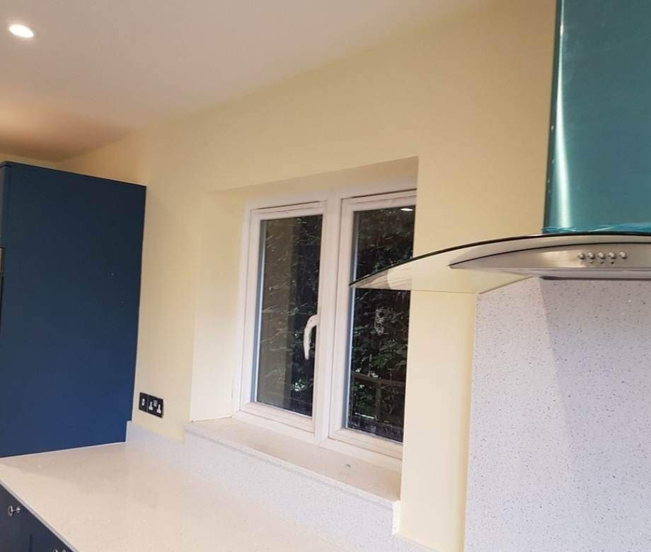 Świeżo pomalowana ściana w kuchni na kremowy kolor, widoczne gniazdka elektryczne, okno z widokiem na zieleń i fragment niebieskiej szafki.