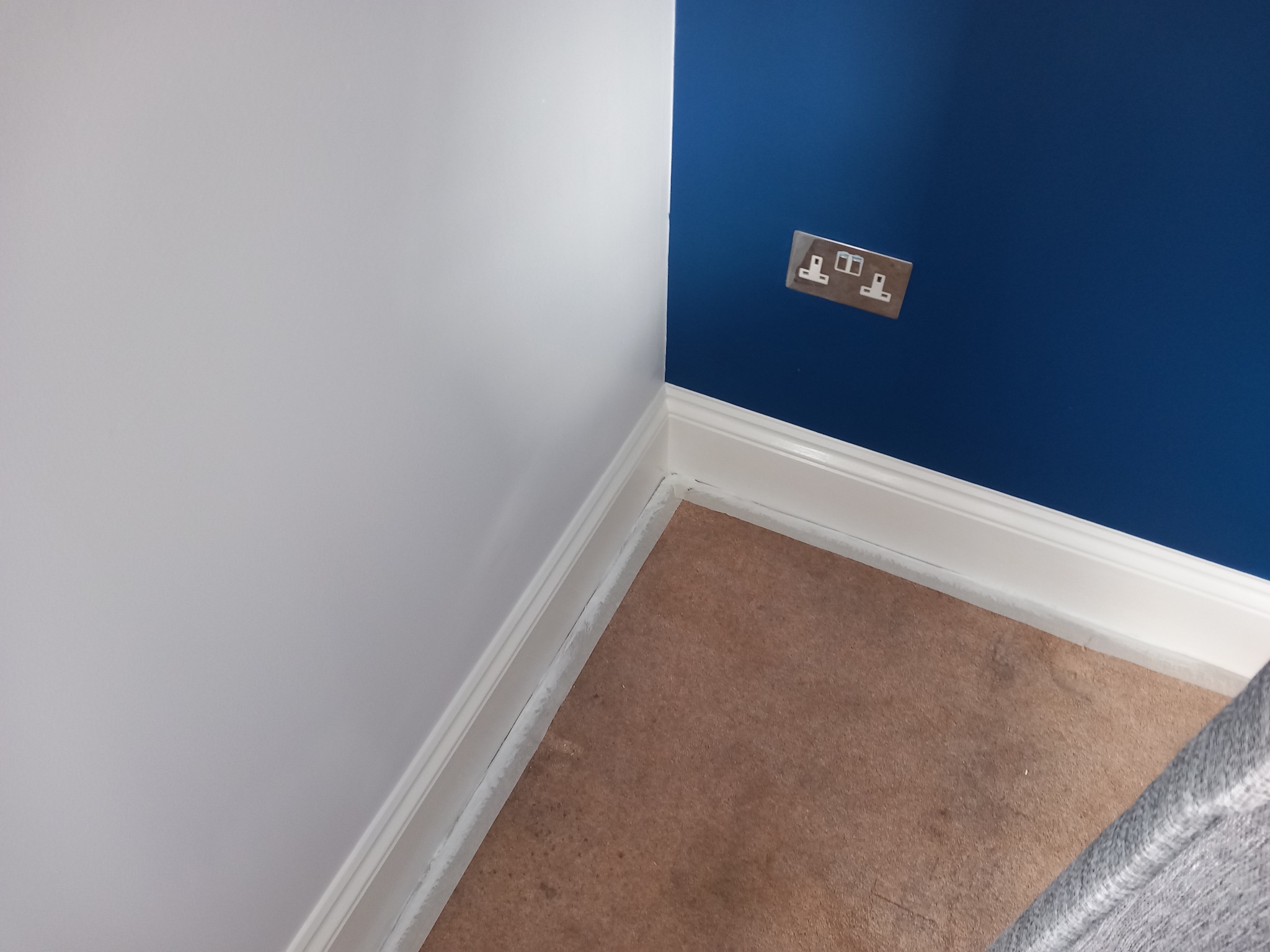 Ściana po malowaniu na dwa kolory (biały i granatowy), z widocznym gniazdkiem elektrycznym i listwą przypodłogową. Widoczne zabezpieczenie podłogi przed zabrudzeniem.