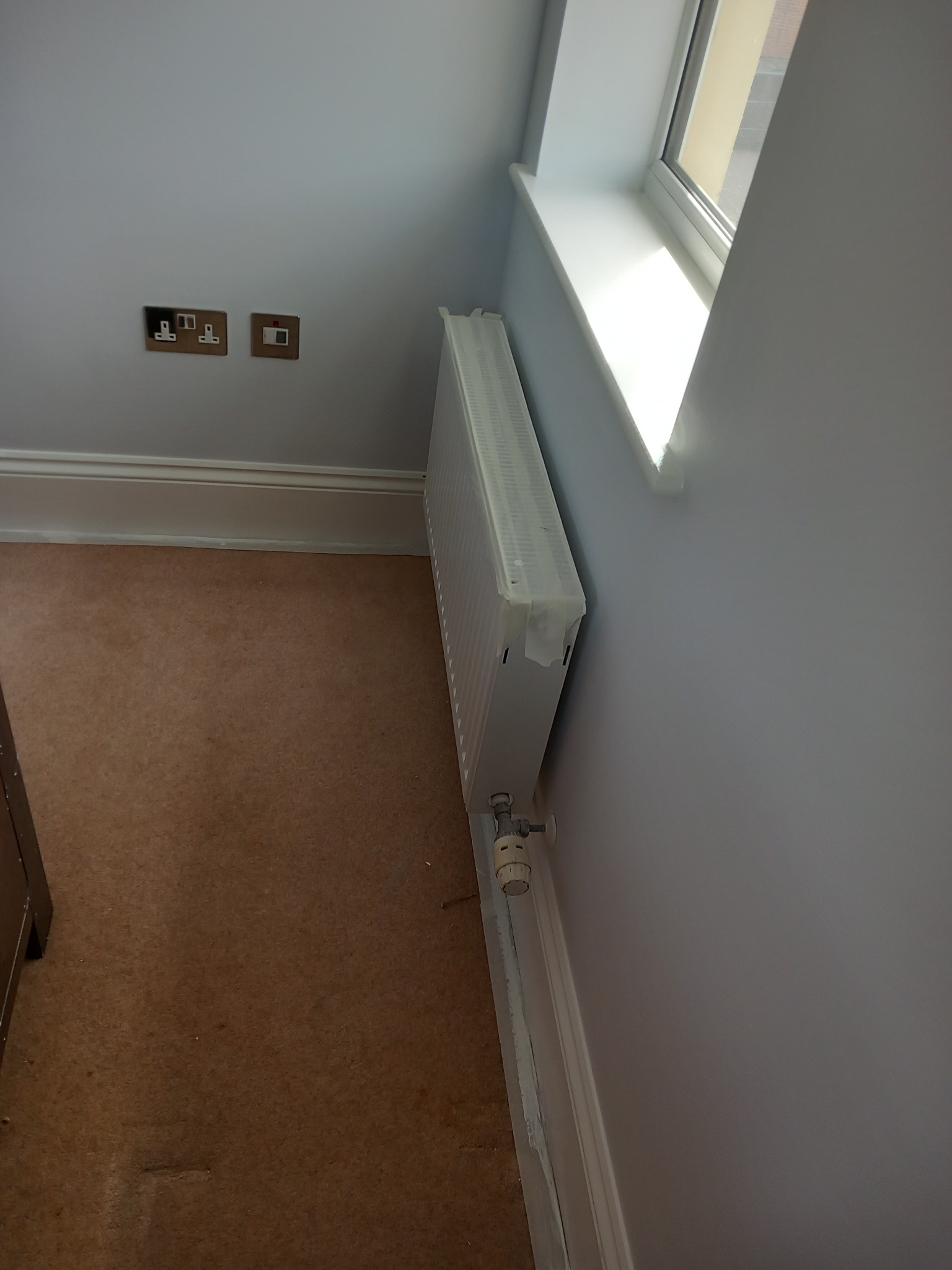 Świeżo pomalowane na szaro wnętrze z grzejnikiem zabezpieczonym taśmą malarską, gładkie ściany, brązowa podłoga i okno z białym parapetem.