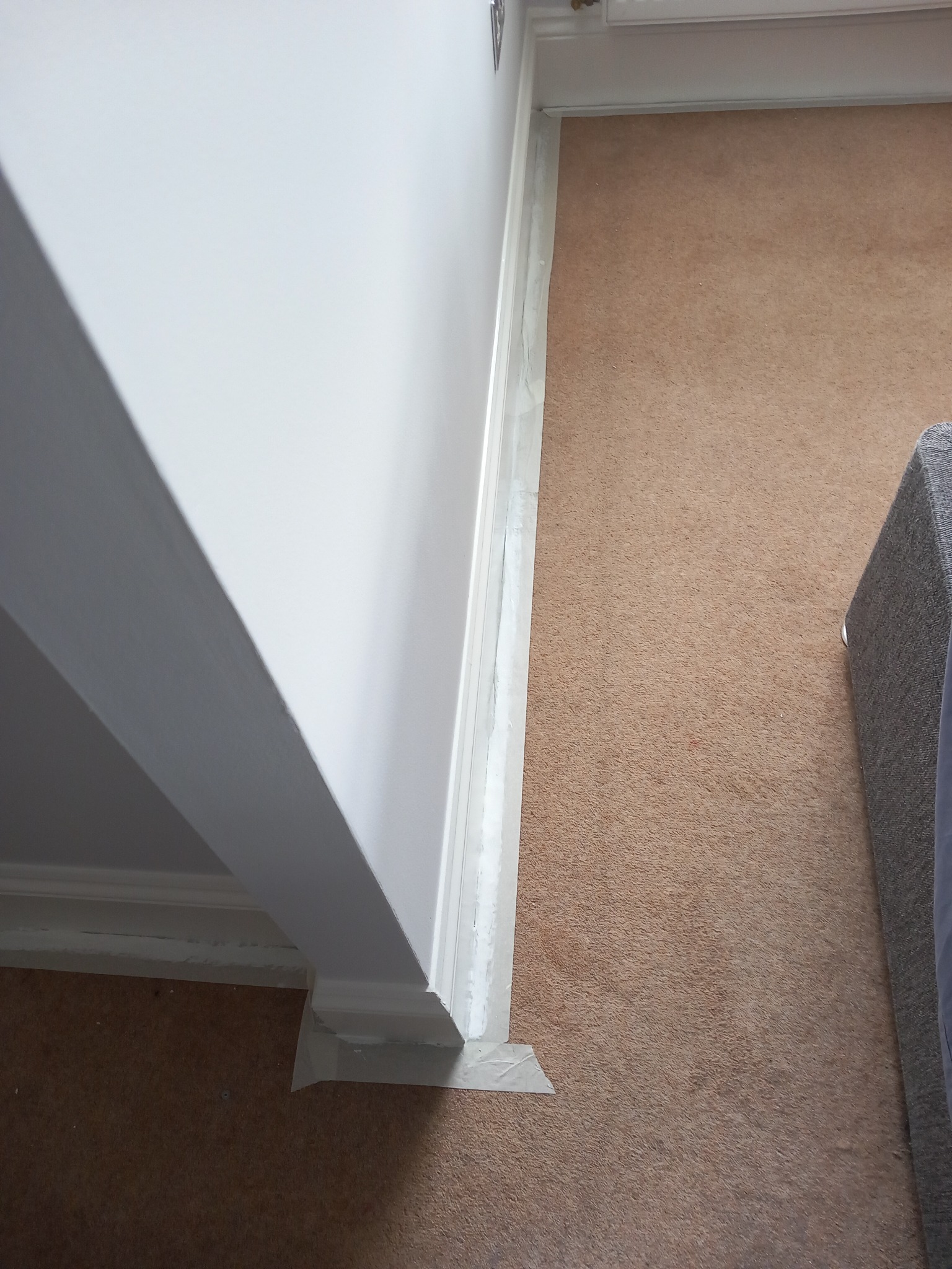 Świeżo pomalowana, biała ściana z listwą przypodłogową zabezpieczoną taśmą malarską, widok z góry na brązową wykładzinę i fragment szarej sofy.
