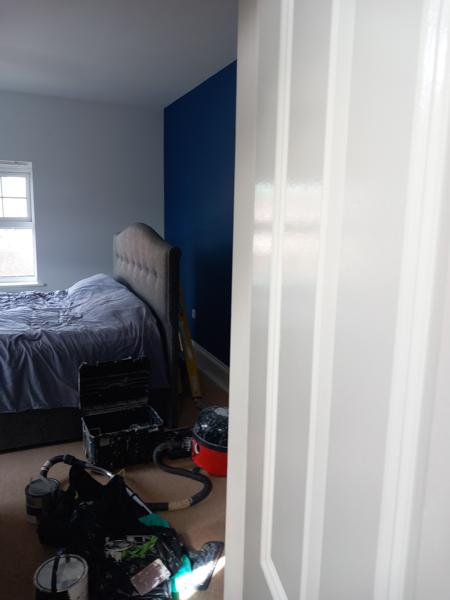 Pokój po malowaniu: granatowa ściana za łóżkiem, narzędzia malarskie na podłodze, widok przez otwarte, białe drzwi.