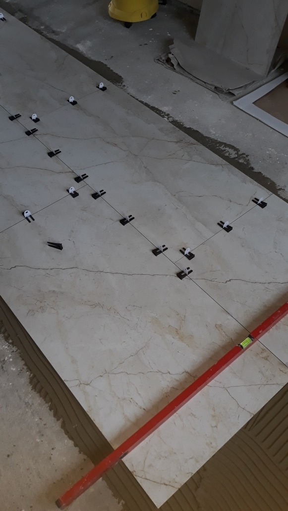 Układanie płytek podłogowych w trakcie realizacji, widoczne systemy poziomowania, poziomica i fragment żółtego kasku ochronnego w tle.