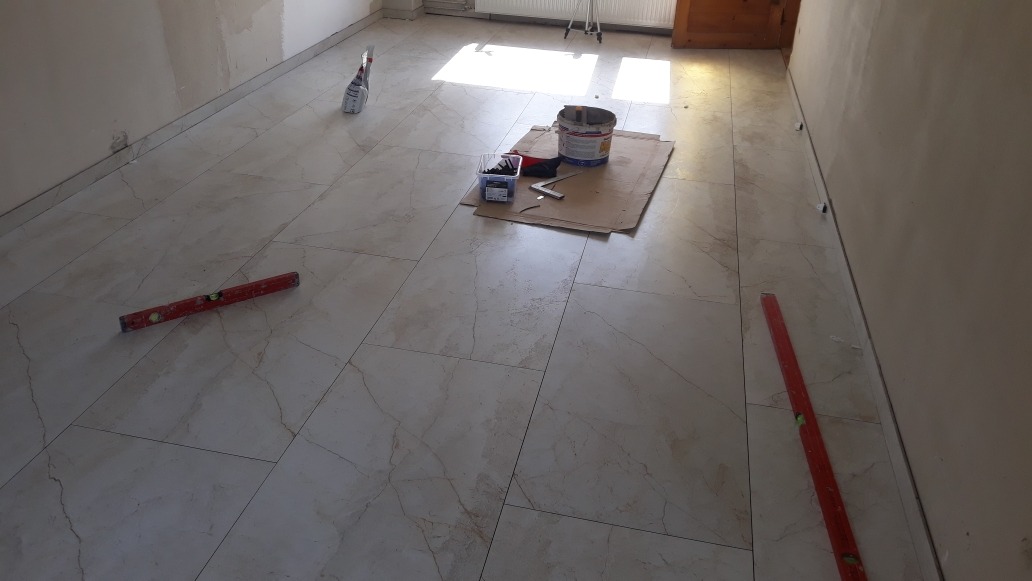 Ułożone marmurowe płytki podłogowe w jasnym pomieszczeniu, poziomica, puszka z klejem, karton ochronny i pudełko z akcesoriami do montażu płytek.