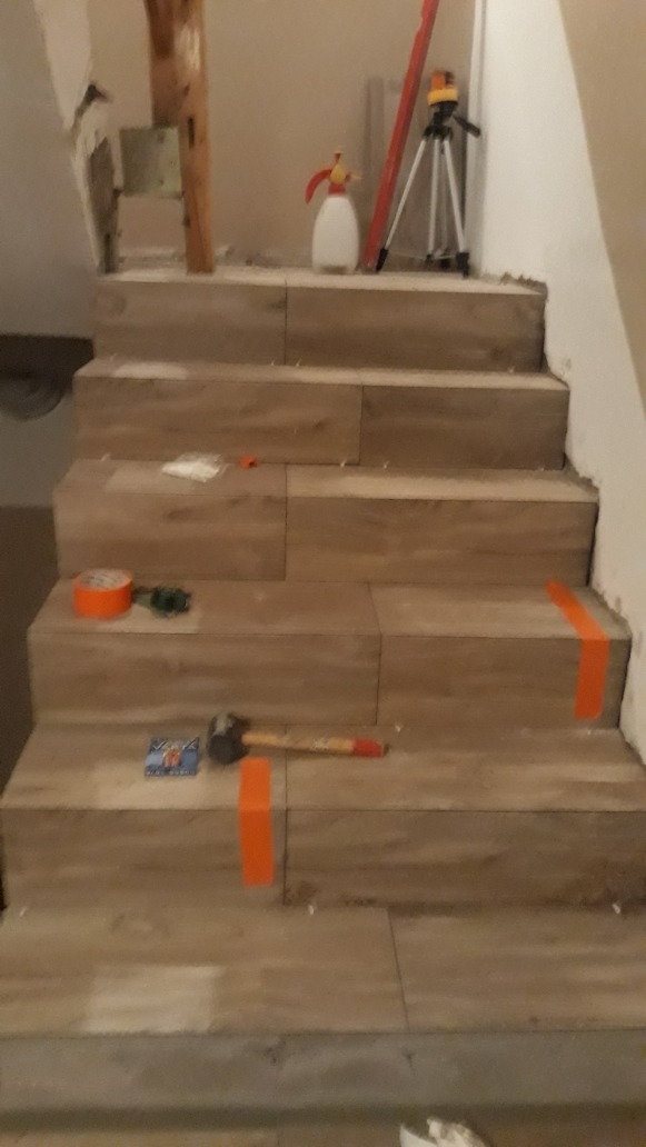 Układanie płytek imitujących drewno na schodach, widoczny młotek, poziomica laserowa na statywie, taśma miernicza i fugi między płytkami.