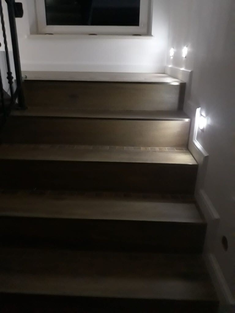 Drewniane schody z oświetleniem punktowym na ścianie, widok z dołu, z fragmentem balustrady po lewej stronie i oknem w tle.