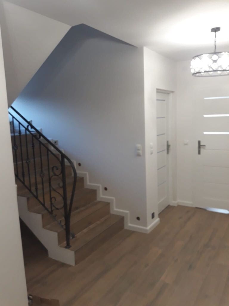 Klatka schodowa w jasnym wnętrzu z drewnianymi stopniami, czarną, ozdobną balustradą i punktowym oświetleniem wzdłuż schodów, widoczna podłoga z paneli, białe drzwi z poziomymi przeszkleniami...