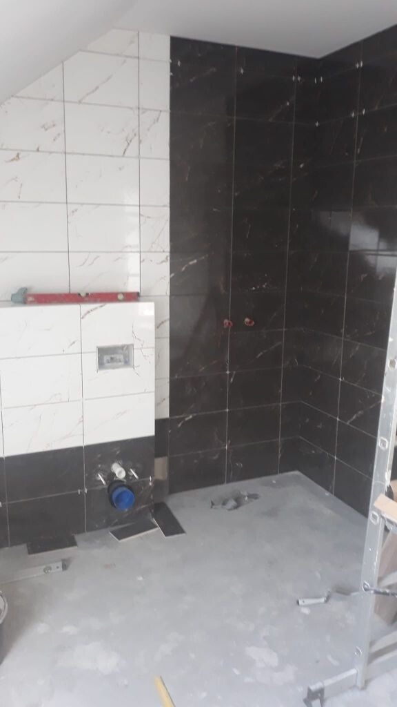 Łazienka w trakcie wykańczania: Połączenie białych i ciemnych płytek imitujących marmur na ścianach, widoczne elementy instalacji podtynkowych, poziomica i drabina w tle, surowy beton na podłodze...