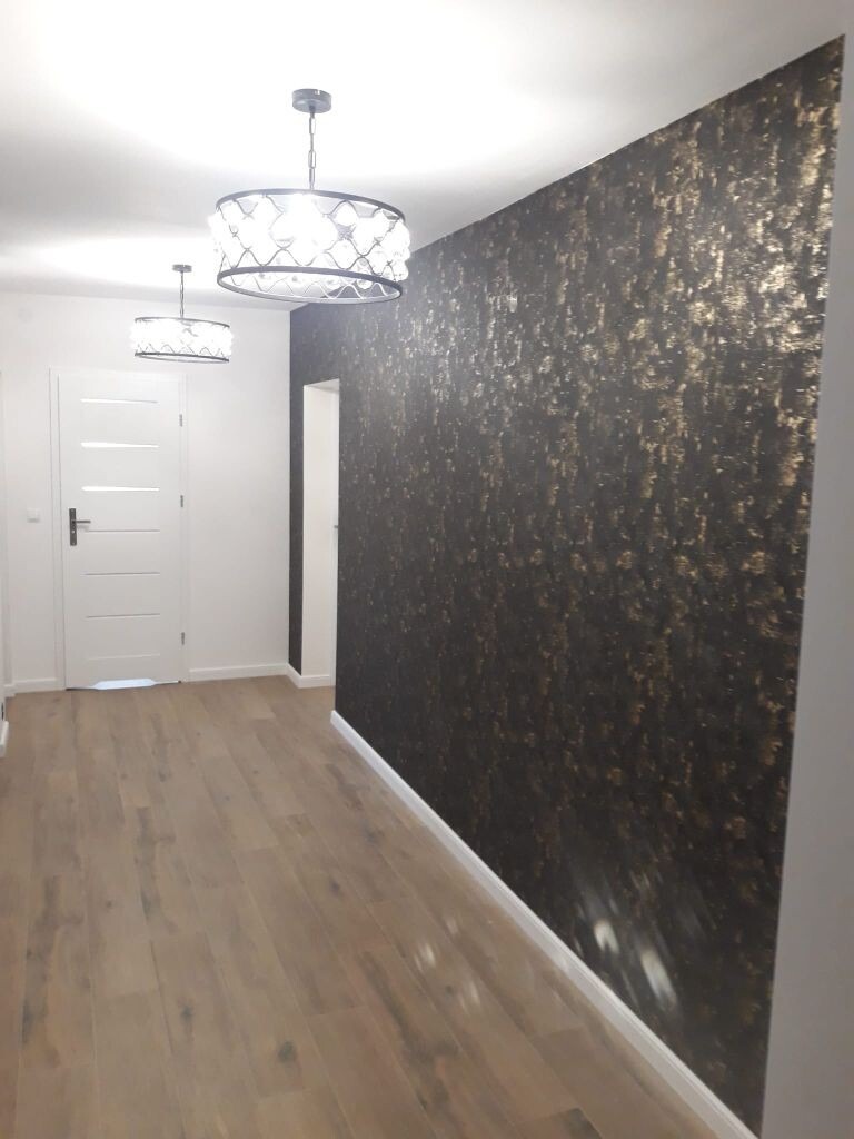 Korytarz z drewnianą podłogą, białą stolarką drzwiową i ścianą pokrytą tapetą w odcieniach czerni i złota, oświetlony dwoma nowoczesnymi lampami sufitowymi.