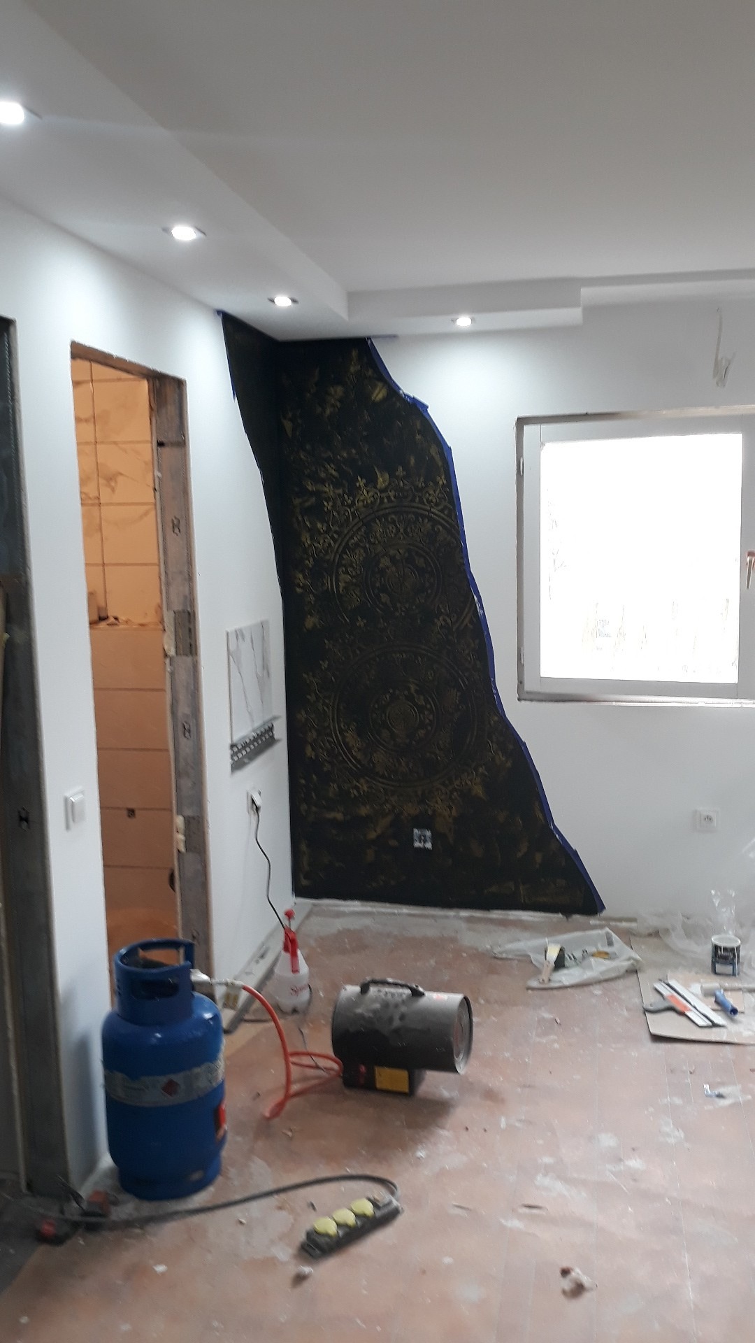 Fragment ściany z dekoracyjną, czarno-złotą strukturą w trakcie tworzenia, widoczny gazowy piecyk grzewczy, butla gazowa i narzędzia malarskie na podłodze.