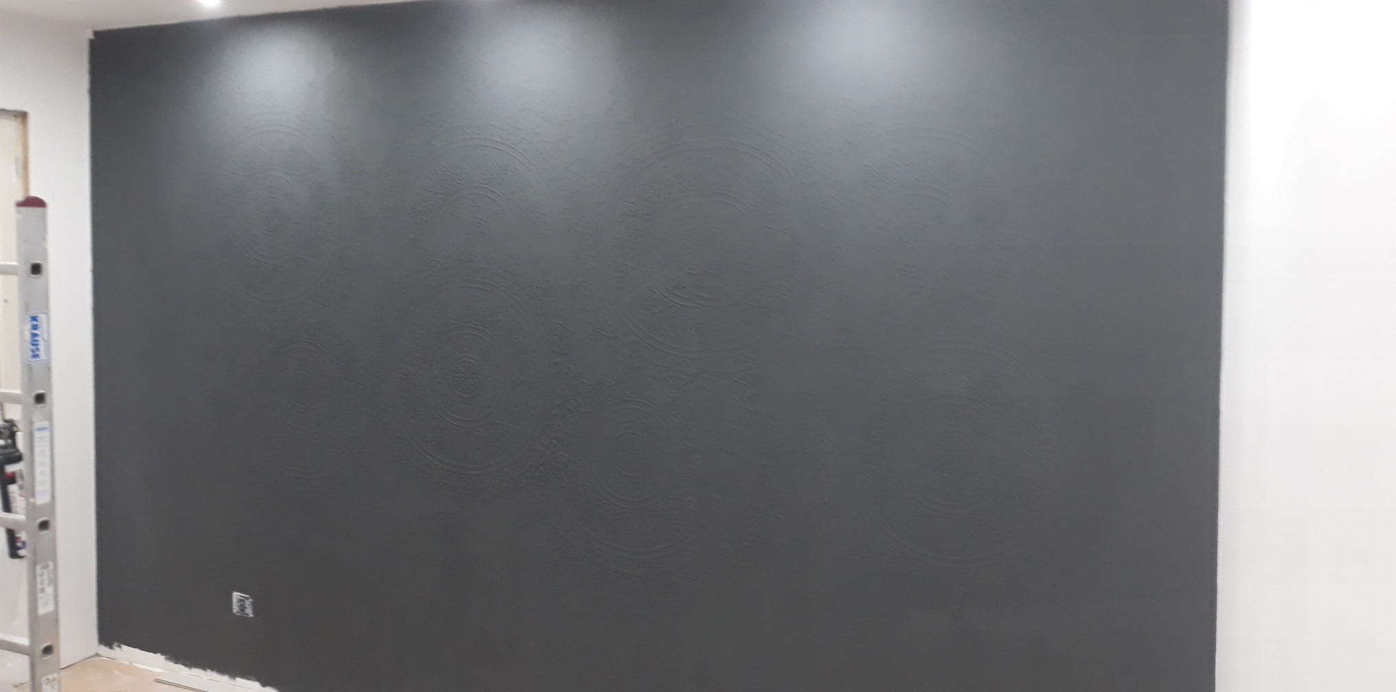 Ściana pomalowana na ciemnoszaro z delikatnym wzorem dekoracyjnym, widoczne ślady po wałku, obok drabina aluminiowa, gniazdko elektryczne na dole ściany.