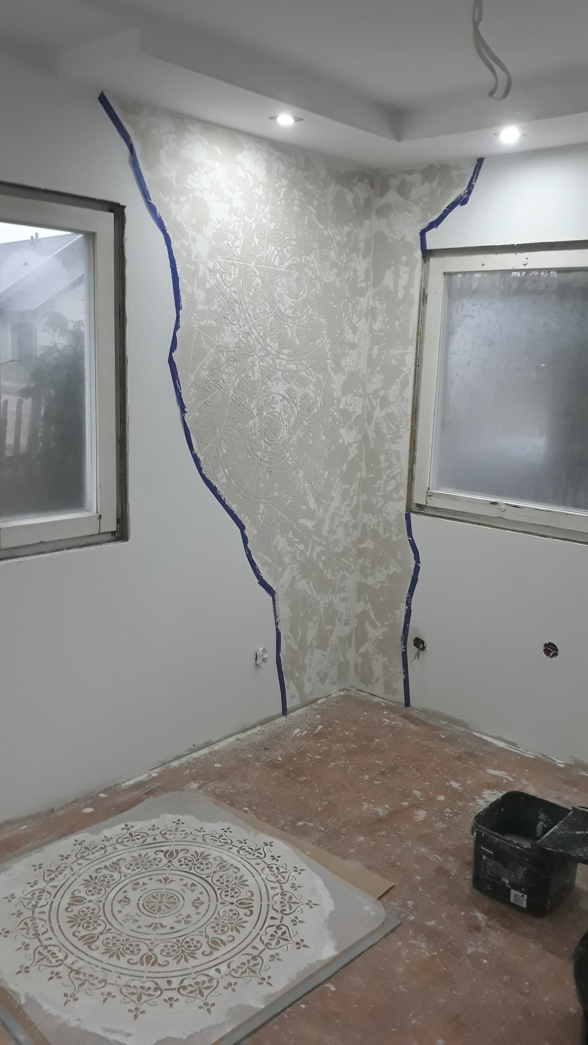 Ściana w trakcie dekoracyjnego wykończenia z użyciem szablonu i taśmy malarskiej, widoczny szablon na podłodze i wiadro z farbą.