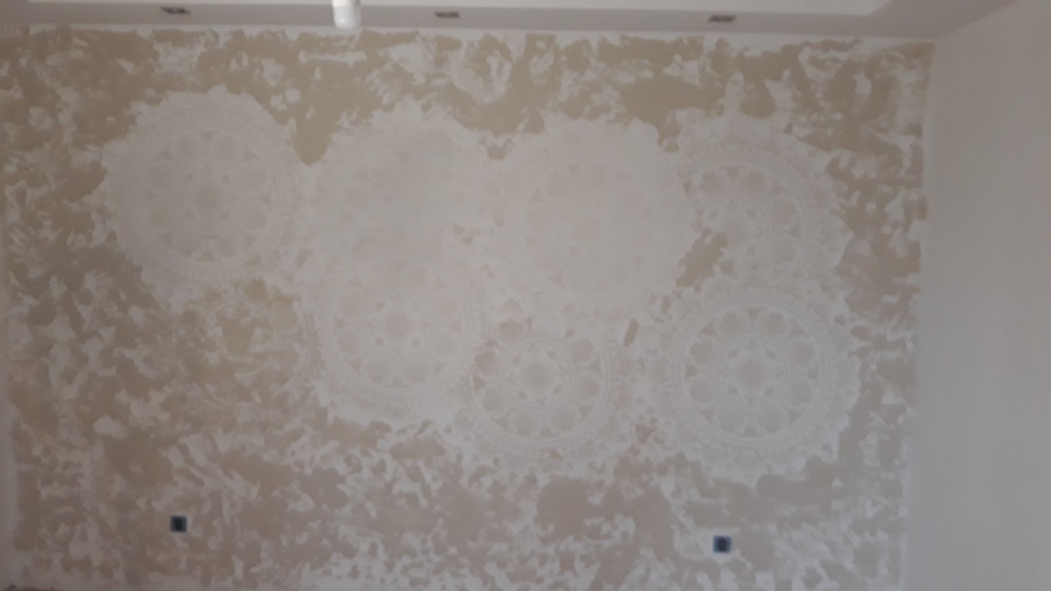 Ściana w beżowych odcieniach z dekoracyjnymi, rozmytymi wzorami mandali, widoczne gniazdka elektryczne