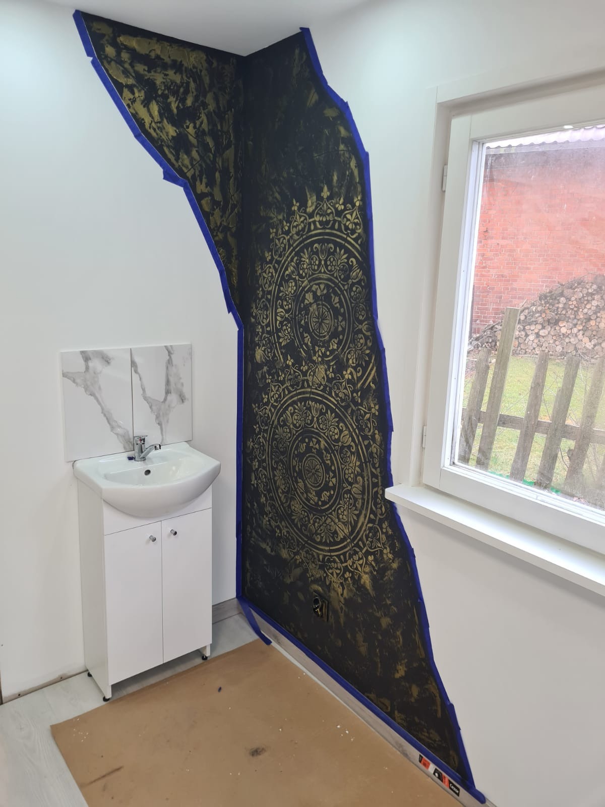 Ściana w łazience z dekoracyjnym, czarno-złotym wzorem, zabezpieczona niebieską taśmą malarską; biała umywalka z szafką i marmurowe płytki nad nią; widok przez okno na podwórko z drewnem.