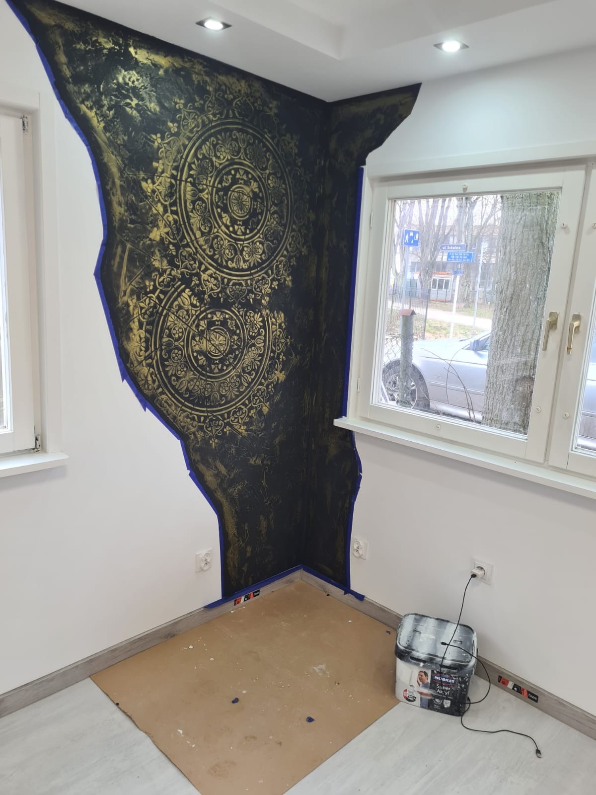 Fragment ściany w trakcie dekorowania techniką szablonową z użyciem farby w kolorze czarnym i złotym, zabezpieczony taśmą malarską, widoczna puszka z farbą.