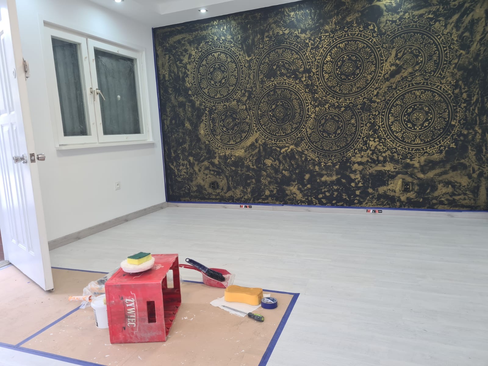 Ściana z dekoracyjnym malowaniem w odcieniach złota i czerni z motywem mandali, zabezpieczona taśmą malarską, widoczne akcesoria malarskie i wiadro na podłodze.