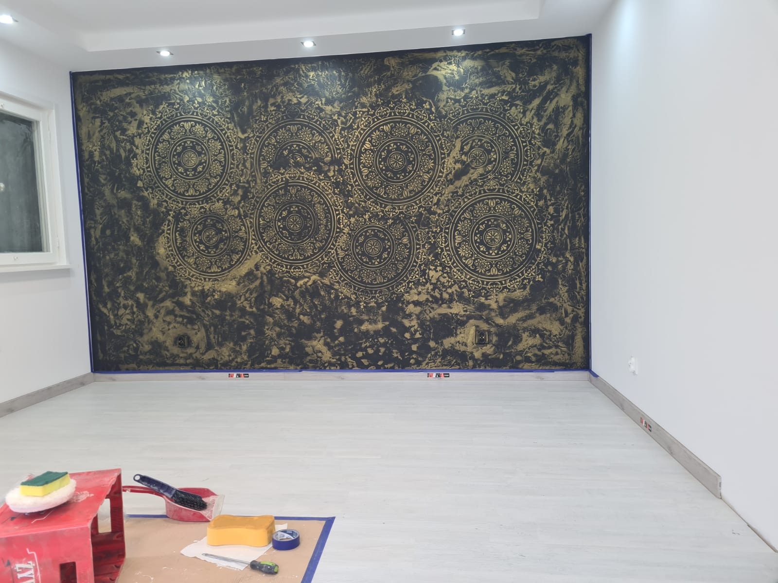 Ściana w pokoju z dekoracyjną, czarno-złotą tapetą z motywem mandali, zabezpieczona niebieską taśmą malarską, z widocznymi narzędziami malarskimi na podłodze.