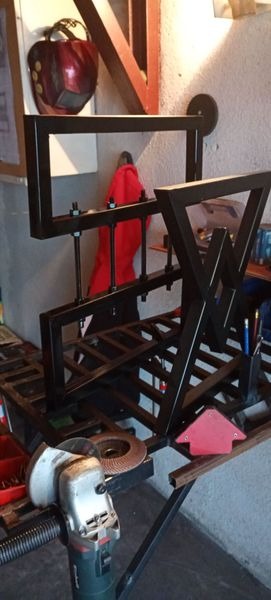 Metalowa konstrukcja spawana z profili, częściowo pomalowana na czarno, w tle maska spawalnicza i czerwony materiał, na pierwszym planie szlifierka kątowa i magnes spawalniczy.