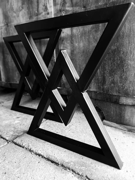 Dwie czarne, metalowe podstawy stołu w kształcie trójkątów, ustawione na betonowej podłodze, sfotografowane w czerni i bieli.