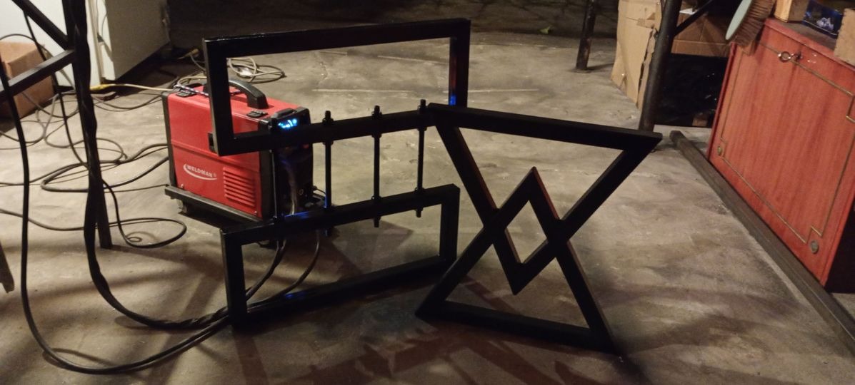 Częściowo zmontowana, czarna, metalowa konstrukcja przypominająca stolik kawowy z motywem górskim, obok czerwona spawarka Weldman z niebieskimi diodami, na betonowej podłodze.