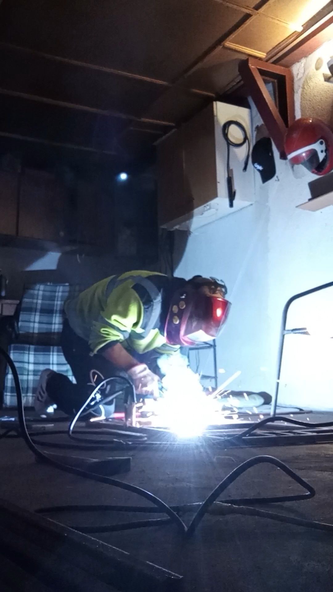 Osoba w masce ochronnej i odblaskowej kamizelce spawa metal w słabo oświetlonym pomieszczeniu, widoczne iskry i kable spawalnicze na podłodze.