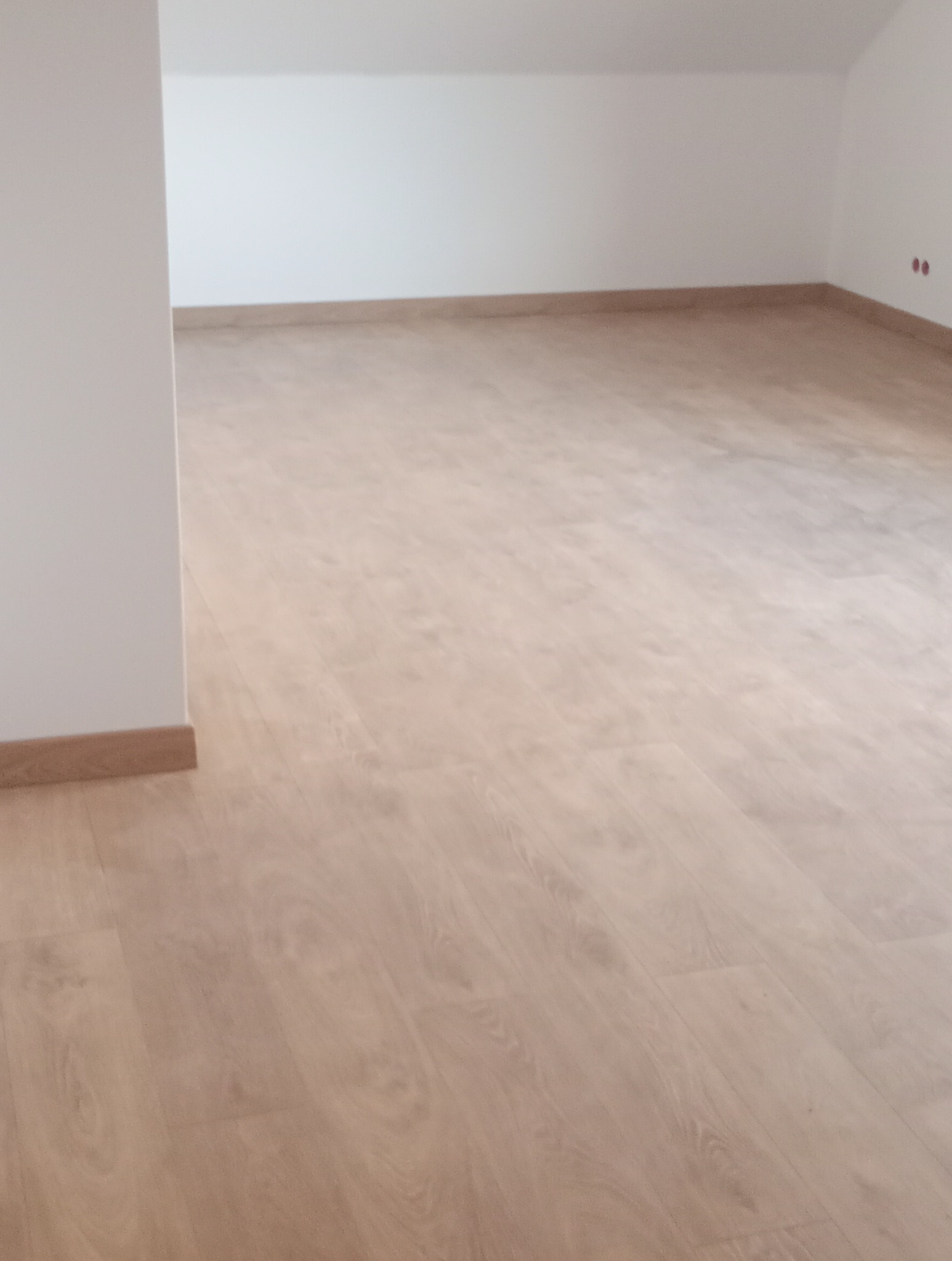 Jasne panele podłogowe ułożone w pustym pomieszczeniu z białymi ścianami i niskim sufitem, widoczne listwy przypodłogowe.