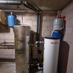 Akj serwis Anna jędral - Dwa zbiorniki buforowe w kotłowni: stalowy i biały z logo Warm, rury instalacyjne pod sufitem z cegły.