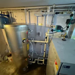 Instalacja w piwnicy z srebrnym zbiornikiem ciepłej wody, pompą obiegową i piecem 21kW, widoczne rury i manometr.