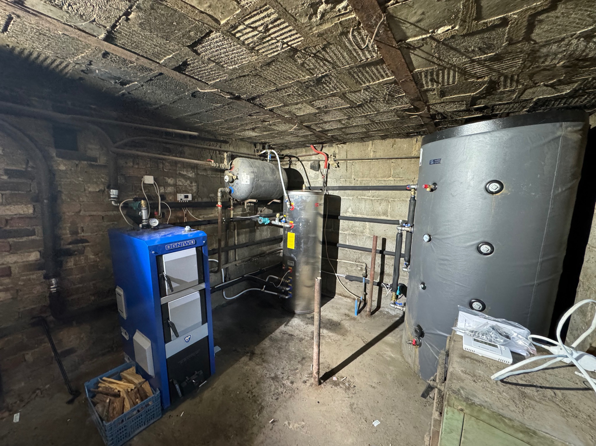 Kocioł na paliwo stałe Ogniwko w kotłowni, obok zbiornik akumulacyjny, rury instalacji grzewczej i wiadro z drewnem opałowym. Na zbiorniku leżą dokumenty i akcesoria montażowe. Widoczny surowy...