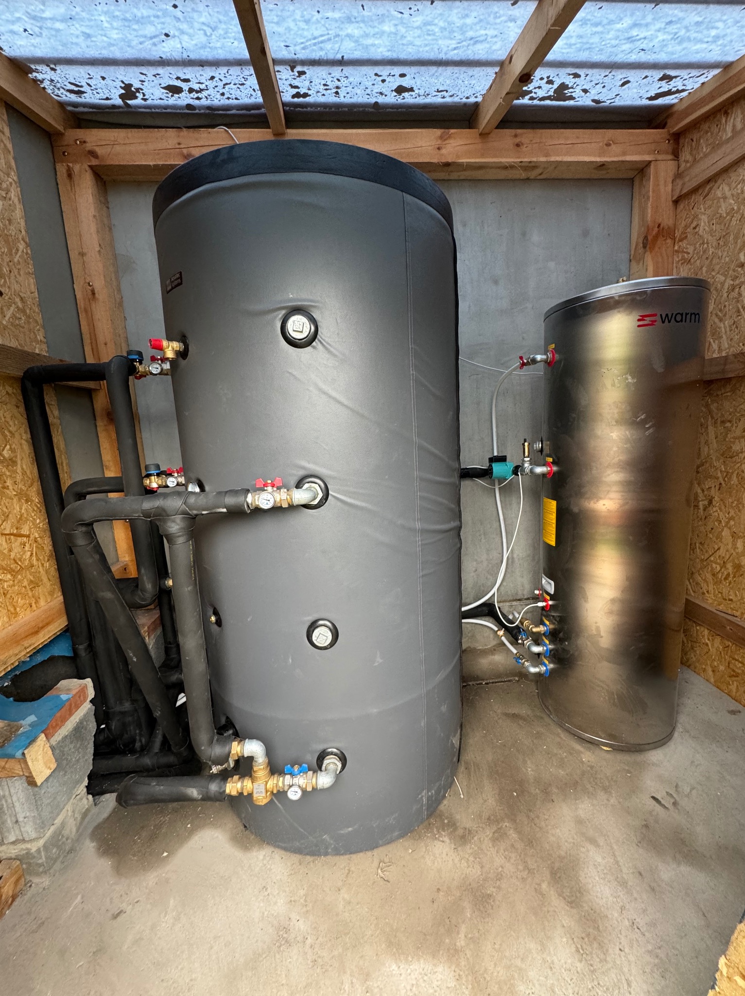 Dwa zbiorniki akumulacyjne: szary, większy z izolacją i czarnymi rurami oraz mniejszy, srebrny z widocznym logo 'warm', zainstalowane w drewnianej konstrukcji z betonową podłogą.