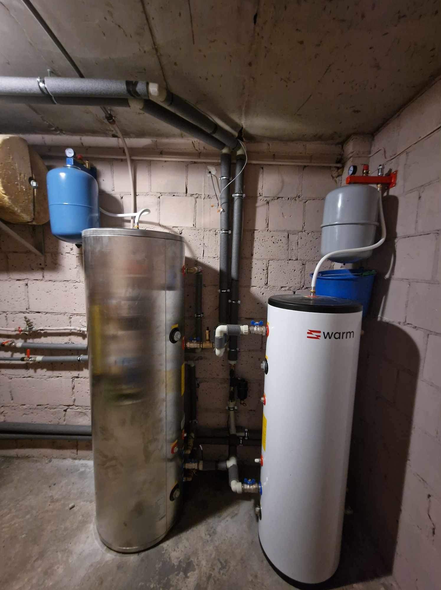Dwa zbiorniki buforowe w kotłowni: stalowy i biały z logo Warm, rury instalacyjne pod sufitem z cegły.
