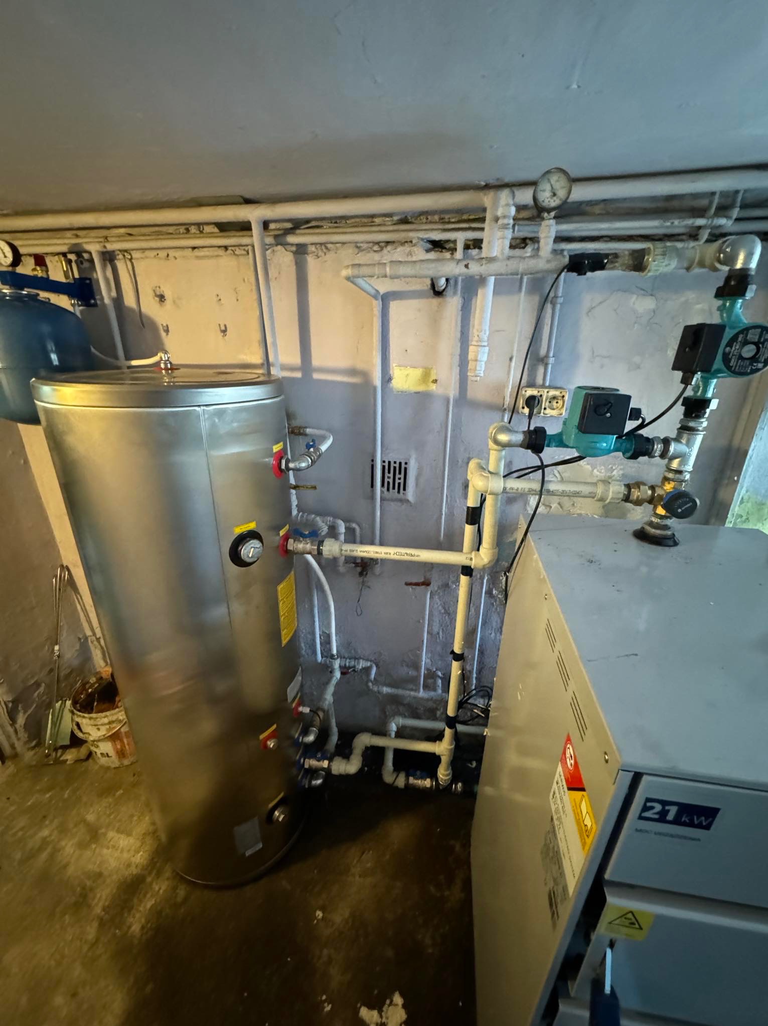 Instalacja w piwnicy z srebrnym zbiornikiem ciepłej wody, pompą obiegową i piecem 21kW, widoczne rury i manometr.