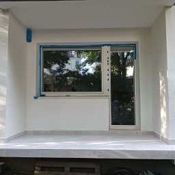 KAMMAR - Wykończony balkon z białymi ścianami o chropowatej strukturze, szarymi płytkami na podłodze i nowymi oknami zabezpieczonymi taśmą malarską.