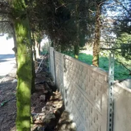 Montaż betonowego ogrodzenia panelowego w terenie leśnym, widoczne słupki montażowe i fragmenty wykopu.