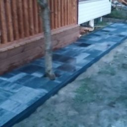 KAMMAR - Ścieżka z szarej kostki brukowej prowadząca od drewnianego tarasu wzdłuż ściany budynku na zewnątrz, obok rosnącego drzewa.