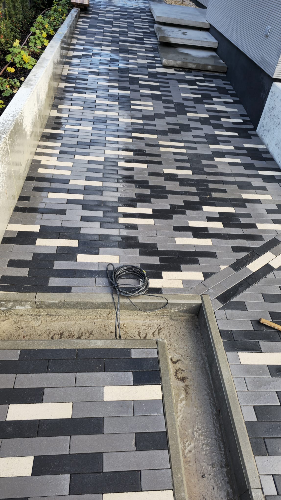 Świeżo ułożona nawierzchnia z prostokątnej kostki brukowej w odcieniach szarości i bieli, widoczny kabel zasilający na krawędzi oraz fragment drewnianego narzędzia, sugerujący trwające prace...