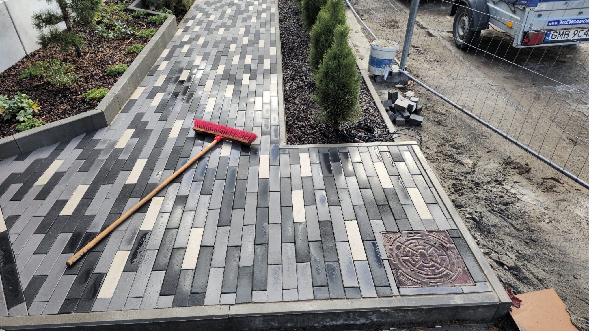 Ułożona ścieżka z kostki brukowej w odcieniach szarości i bieli, z czerwoną miotłą opartą o powierzchnię. Widoczny fragment trawnika z niskimi drzewkami i studzienka kanalizacyjna.