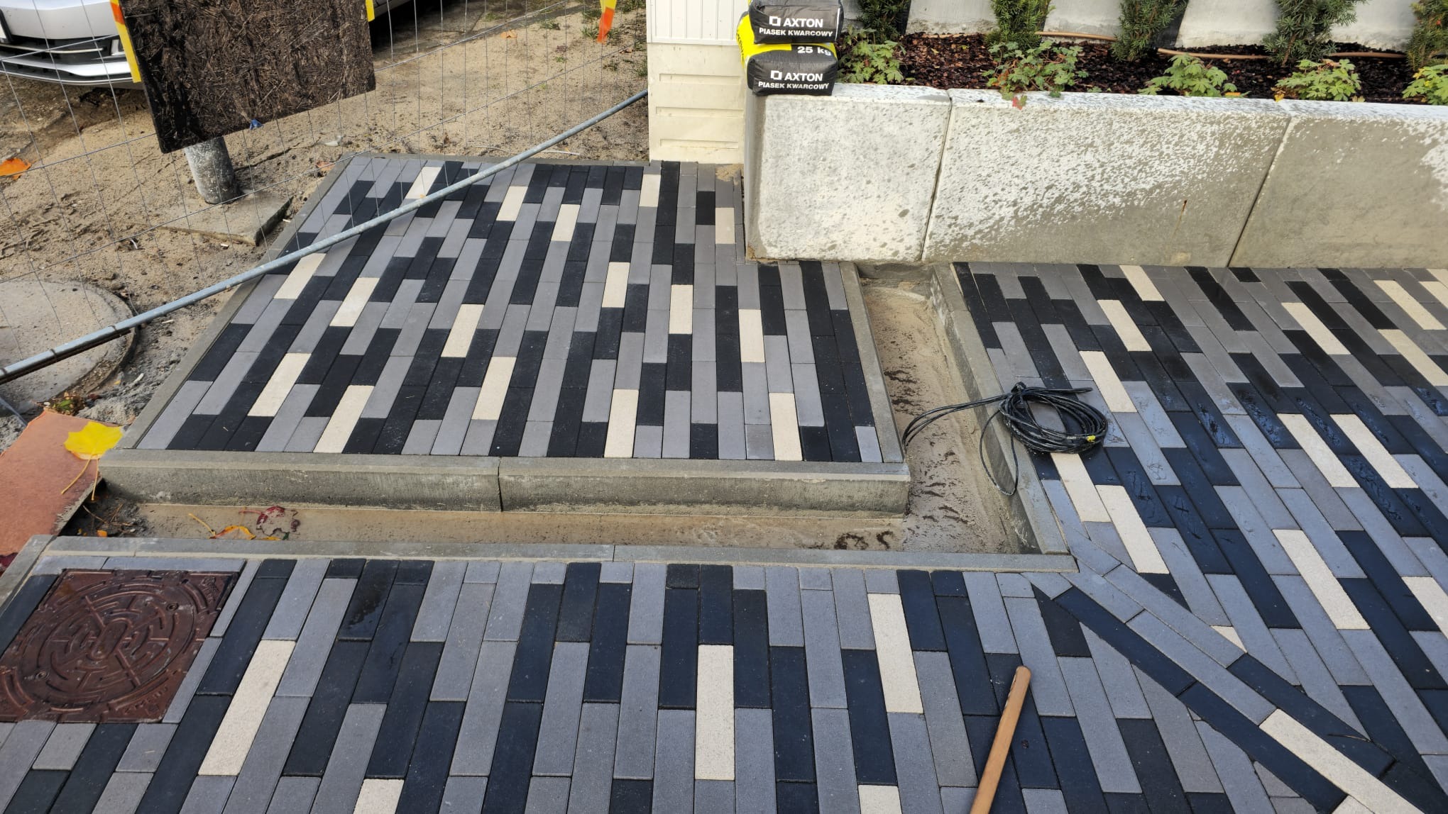 Fragment ścieżki z kostki brukowej ułożonej w regularny wzór z elementów w odcieniach szarości, czerni i beżu, widoczne dwa worki piasku kwarcowego Axton, kabel elektryczny i studzienka kanalizacyjna