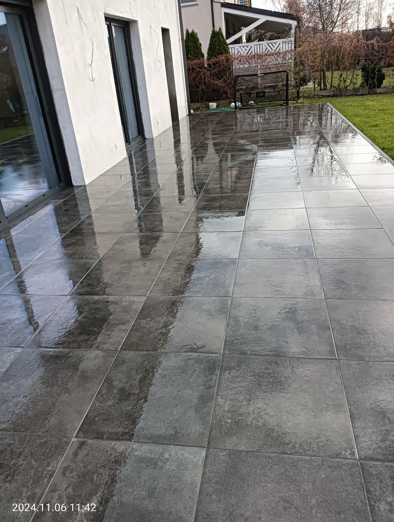 Świeżo ułożona, mokra, ciemnoszara kostka brukowa na tarasie przydomowym, odbijająca światło i architekturę budynku; widoczny fragment trawnika i ogrodzenia w tle.