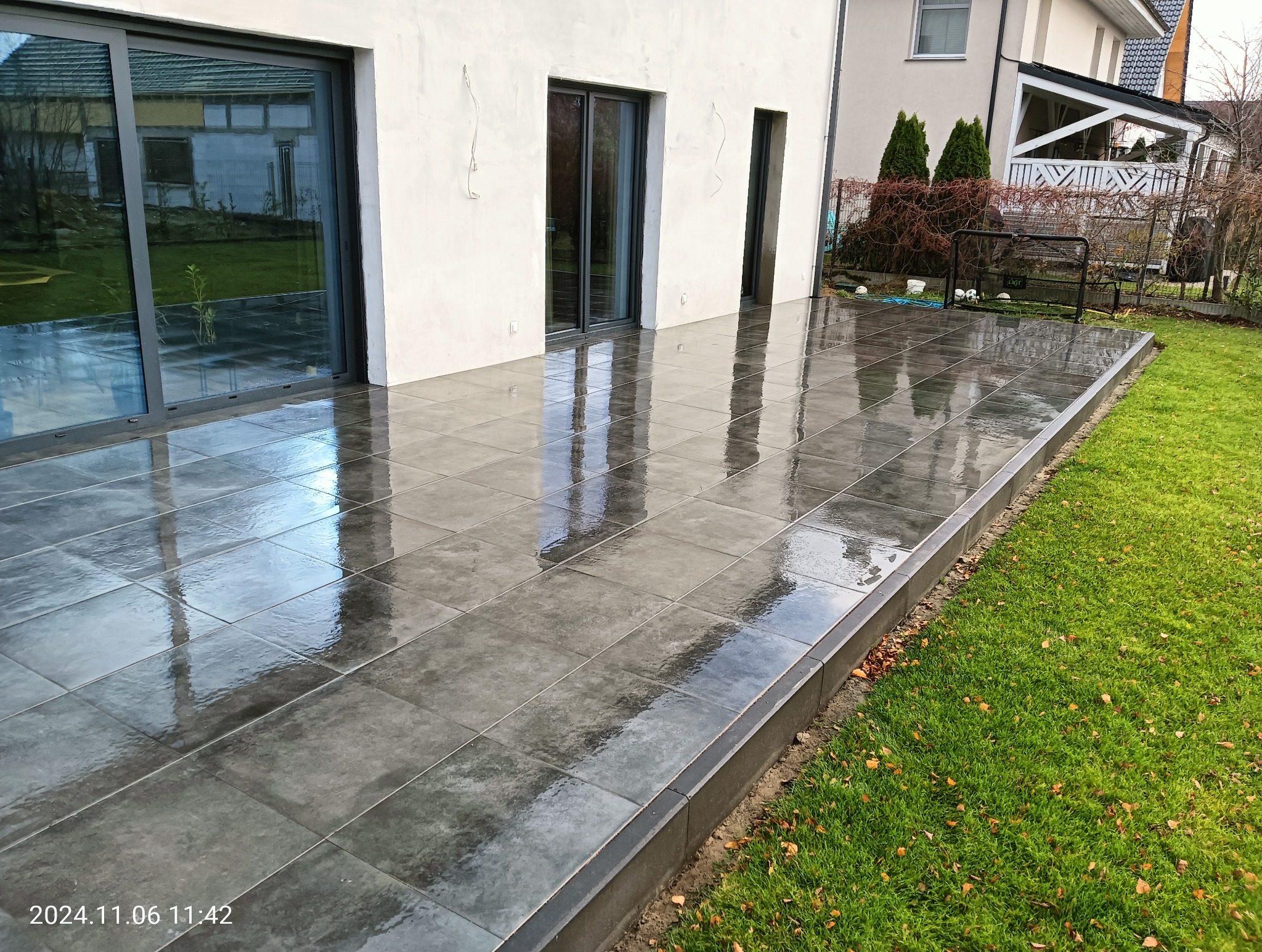 Świeżo ułożony, mokry taras z szarych, kwadratowych płytek, przylegający do białego budynku z dużymi przesuwnymi drzwiami, obrzeża z ciemnego betonu i trawnik z opadłymi liśćmi na obrzeżach.