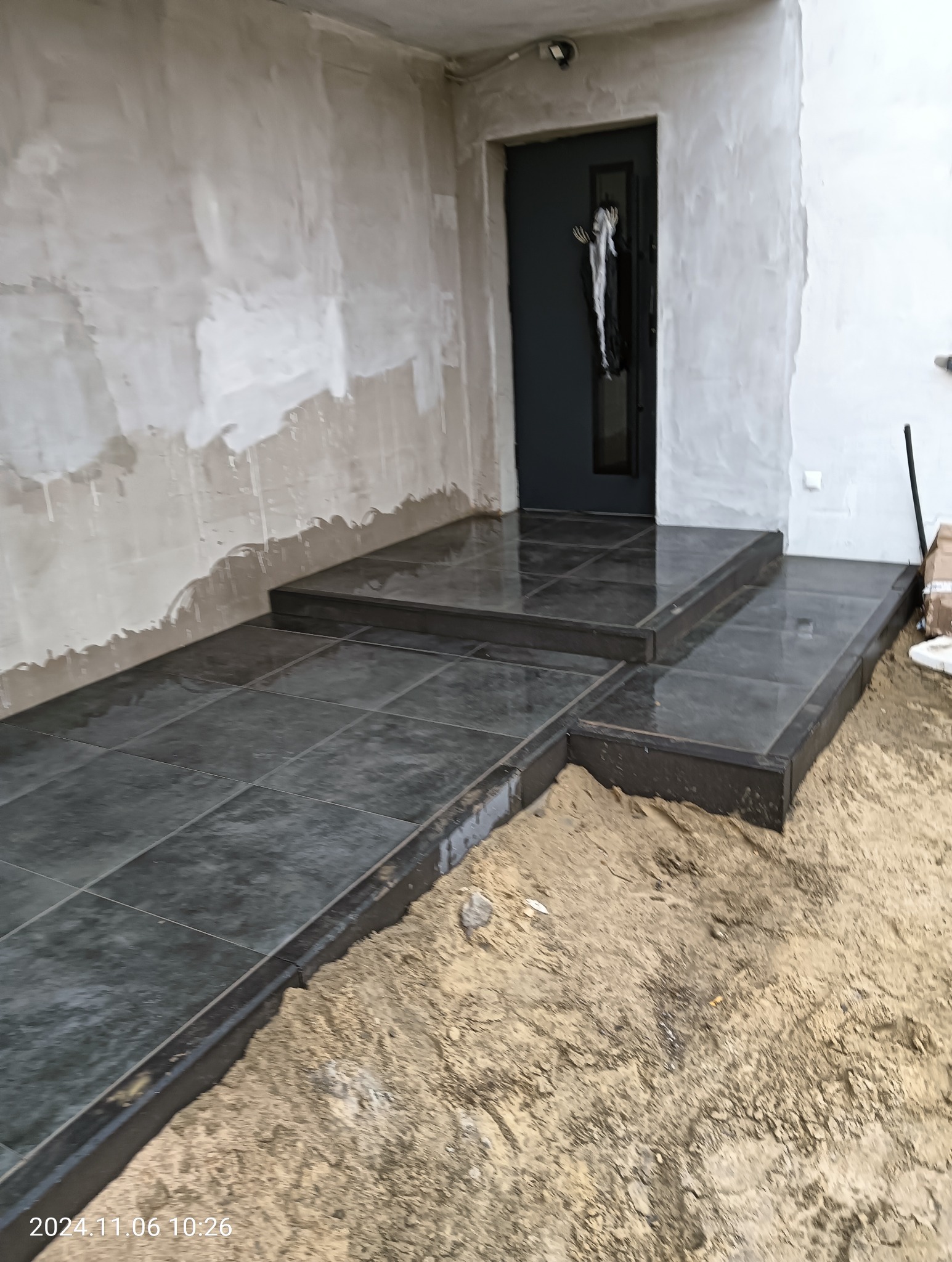Ułożone, ciemnoszare płytki na schodach wejściowych do domu, z widoczną surową ścianą i piaskiem wokół, wskazujące na trwające prace wykończeniowe.